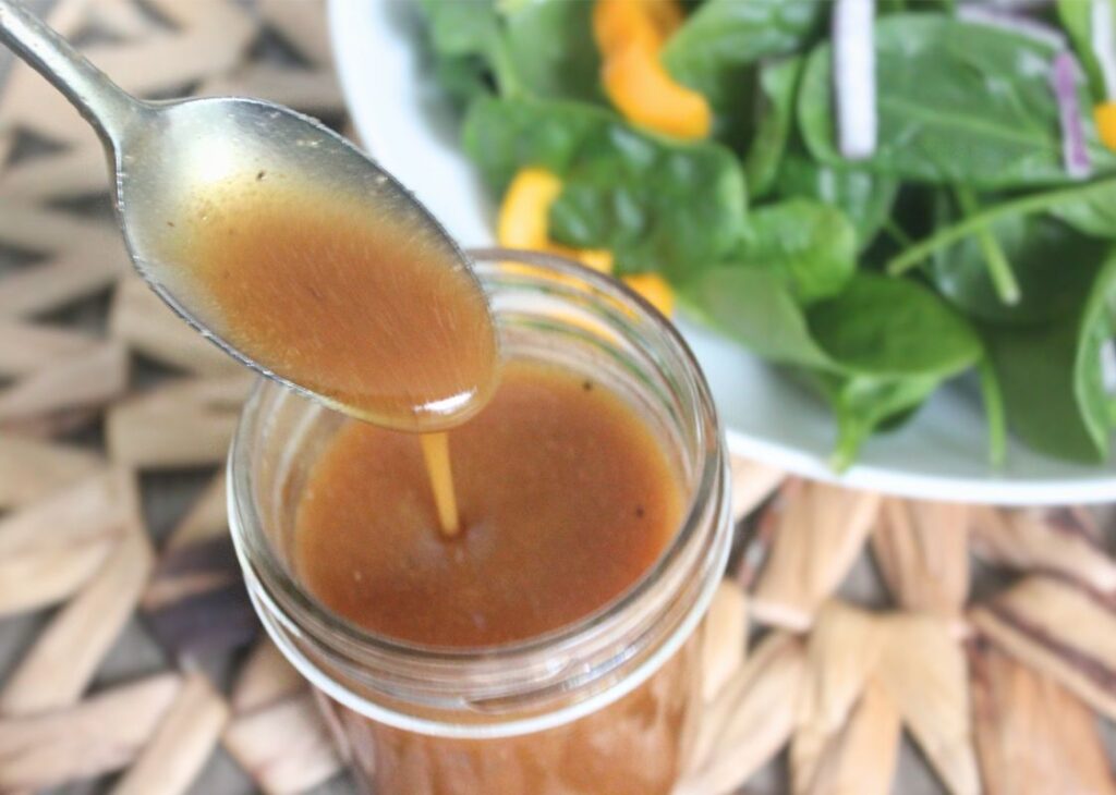 Honey ginger salad dressing being poured from a spoon into a glass jar with fresh spinach salad in the background.