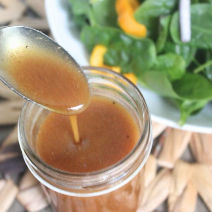 Honey ginger salad dressing being poured from a spoon into a glass jar with fresh spinach salad in the background.