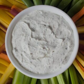 White bowl of dairy free vegetable dip surrounded by carrots, celery, and snap peas fanned out on a white plate.