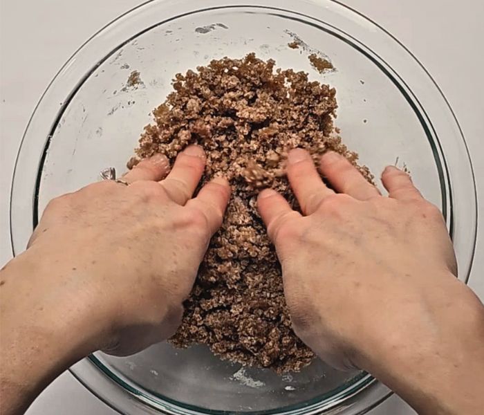 Hands pressing crumb topping mixture together in a glass bowl to form large crumbs