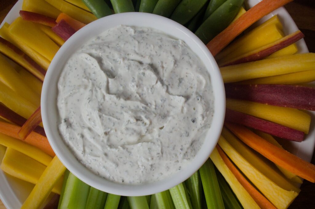 A white bowl of creamy vegetable dip surrounded by colorful vegetable crudités including yellow and orange carrots, celery and cucumber arranged in a fan pattern on a white plate