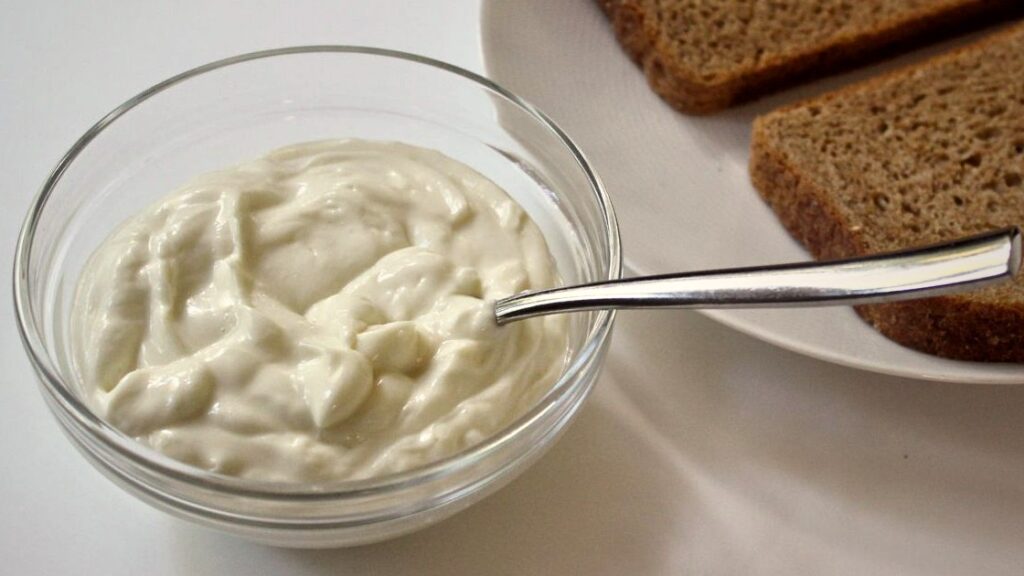 Glass bowl of homemade avocado oil mayonnaise with a spoon on a white surface next to slices of bread