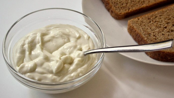mustard-free-mayonnaise-recipe-700 Glass bowl of homemade avocado oil mayonnaise with a spoon on a white surface next to slices of bread