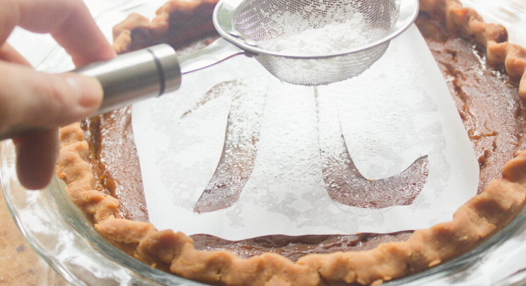 A hand holds a fine mesh sifter dusting powdered sugar through a pi symbol stencil onto a pumpkin pie in a glass pie dish