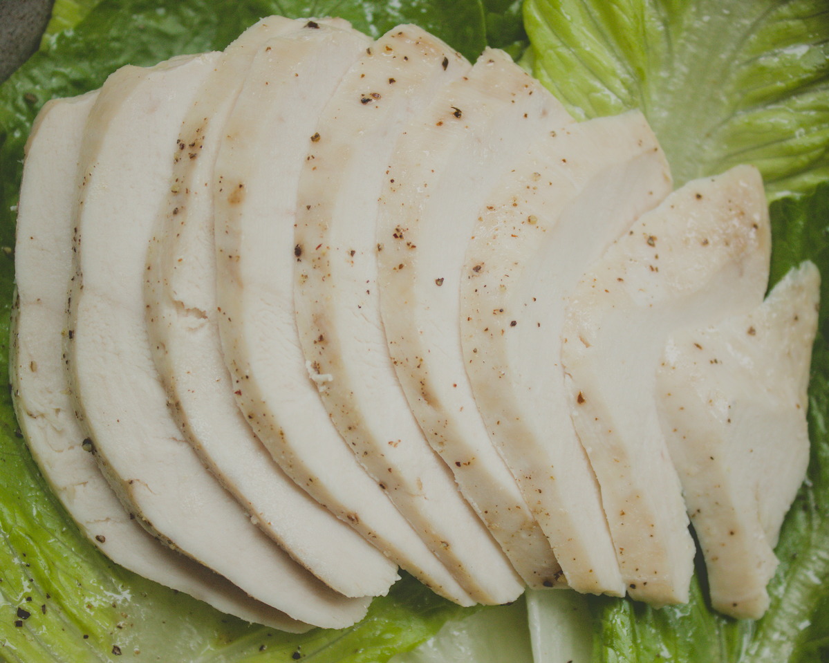Thinly sliced cooked chicken breast arranged in a fan pattern on a bed of fresh green romaine lettuce leaves, seasoned with black pepper