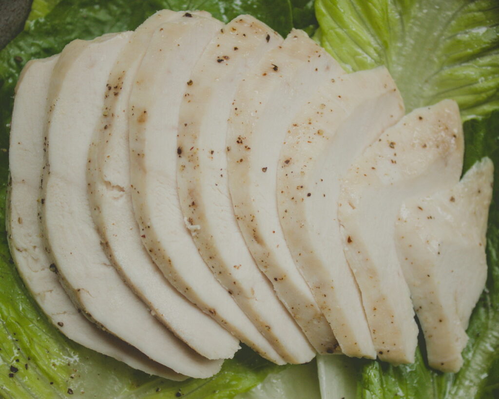 Thinly sliced cooked chicken breast arranged in a fan pattern on a bed of fresh green romaine lettuce leaves, seasoned with black pepper
