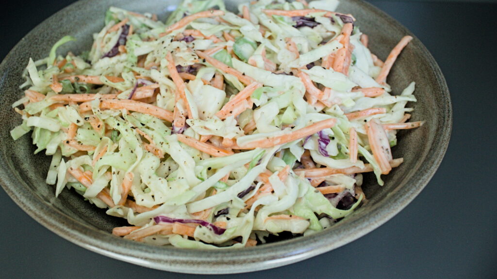 A bowl of fresh homemade coleslaw with shredded green cabbage, carrots, and red cabbage, served in a ceramic bowl on a dark background