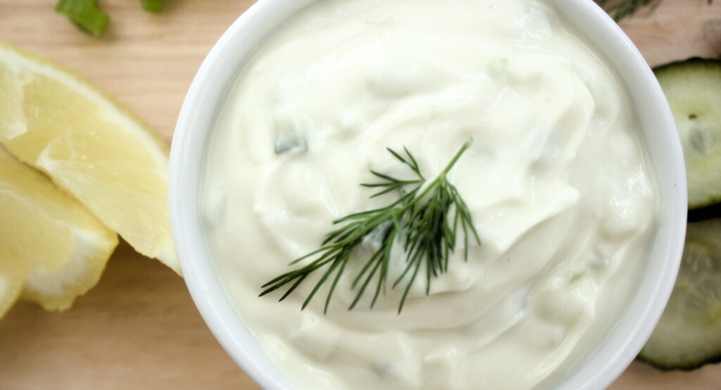 Creamy homemade tartar sauce in a white ramekin topped with fresh dill, served with lemon wedges and cucumber slices on a wooden board