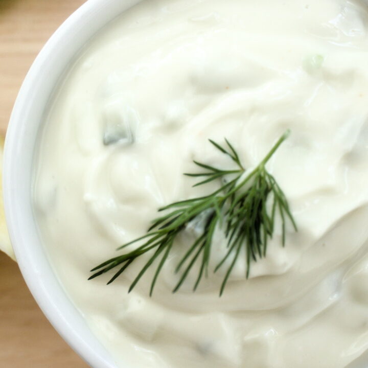 Creamy homemade tartar sauce in a white ramekin topped with fresh dill, served with lemon wedges and cucumber slices on a wooden board